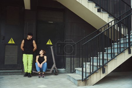 Foto de Pareja moderna de informales está descansando cerca de la pared del edificio de la ciudad, una joven hembra se sentó en un scooter - Imagen libre de derechos