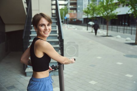 Foto de Sonriente joven mujer de la ciudad se para con un scooter cerca de las escaleras, ella está en ropa deportiva cómoda - Imagen libre de derechos
