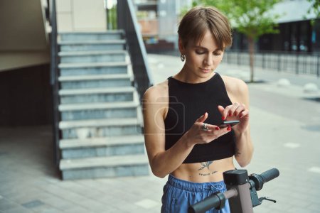 Téléchargez les photos : Jeune femme de la ville se tient avec un scooter et un téléphone dans une rue de la ville, elle utilise une application en ligne - en image libre de droit