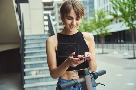 Téléchargez les photos : Moderne jeune femme de la ville se tient avec un scooter et un téléphone sur une rue de la ville - en image libre de droit