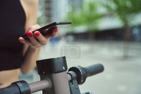 Photo for Young city woman with a scooter and a phone on a city street, she uses an online application - Royalty Free Image