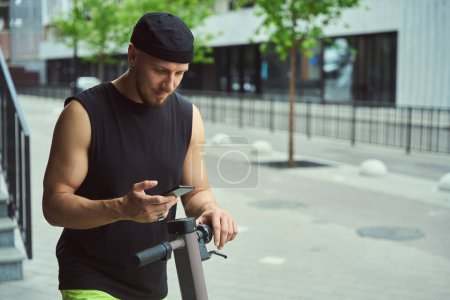 Téléchargez les photos : Jeune homme de la ville avec un scooter et un téléphone dans une rue de la ville, il utilise une application en ligne - en image libre de droit