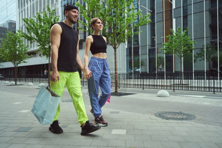 Foto de Un par de informales caminan de la mano, contra el telón de fondo de un edificio moderno, tienen bolsas de compras - Imagen libre de derechos