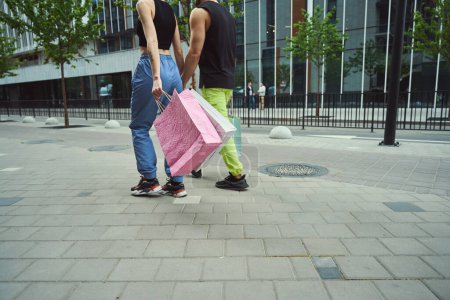Téléchargez les photos : Jeune couple marche main dans la main le long du trottoir, les gens ont des sacs à provisions - en image libre de droit
