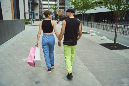 Téléchargez les photos : Guy et sa copine marchent main dans la main le long du trottoir, la femelle a des sacs à provisions - en image libre de droit