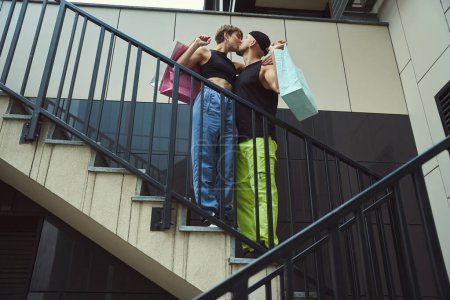 Téléchargez les photos : Guy et sa copine s'embrassent sur les marches des escaliers de la ville, ils ont des sacs à provisions dans leurs mains - en image libre de droit