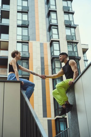 Photo for Young man and his girlfriend are sitting on a high curb, holding hands, next to large modern buildings - Royalty Free Image