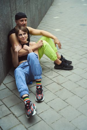 Téléchargez les photos : Jeune homme informel et sa petite amie sont assis, s'embrassant, sur le trottoir dans une petite cour - en image libre de droit