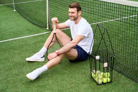 Foto de Sonriente hombre guapo sentado en el césped en la cancha de tenis, tiene una raqueta y una canasta de bolas - Imagen libre de derechos