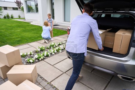 Téléchargez les photos : L'homme porte des boîtes en carton avec des choses d'une voiture à une nouvelle maison, sa femme et sa fille sont à proximité - en image libre de droit