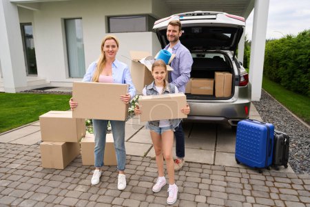 Téléchargez les photos : Heureuse famille se tient avec des boîtes en carton près de la voiture, ils déménagent dans une nouvelle maison - en image libre de droit