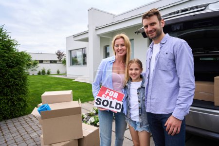 Téléchargez les photos : Les parents avec leur fille se tiennent dans la cour d'une nouvelle maison, une femme tient un signe à vendre - en image libre de droit