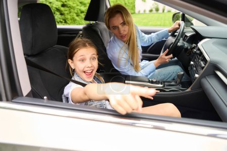 Téléchargez les photos : Joyeux petite fille pointe quelque chose à l'extérieur de la fenêtre de la voiture, sa charmante mère conduit - en image libre de droit