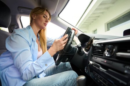Téléchargez les photos : Jeune femme assise au volant d'une voiture moderne, elle a un téléphone portable dans ses mains - en image libre de droit