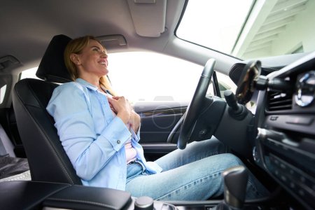 Photo for Young happy woman is sitting behind the wheel of a modern car, she is wearing comfortable casual clothes - Royalty Free Image