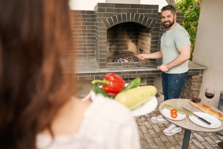Foto de Hombre en jeans construye un fuego en un gran horno en el patio trasero, su esposa le trae verduras - Imagen libre de derechos