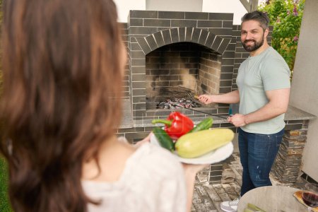 Foto de Hombre en jeans hace un fuego en una gran estufa en el patio trasero, su esposa le trae verduras - Imagen libre de derechos