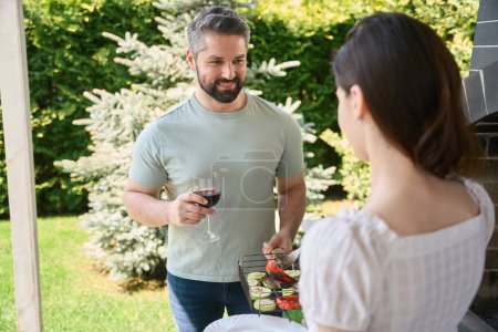 Téléchargez les photos : L'homme tient un gril avec des légumes et un verre de vin, il communique avec sa femme dans la cour arrière - en image libre de droit