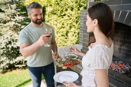 Téléchargez les photos : Femme brune met des légumes grillés dans une assiette, à côté de son mari avec un verre de vin - en image libre de droit
