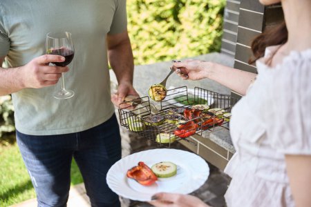 Téléchargez les photos : Femme met des légumes grillés dans son assiette, à côté de son mari avec un verre de vin - en image libre de droit