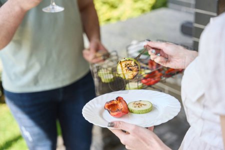 Téléchargez les photos : Femme se tient debout avec une assiette de légumes grillés dans la cour, à côté de son mari avec un verre de vin - en image libre de droit