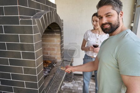 Photo for Male in jeans cooks food in a large oven in the backyard, his wife is next to him with a glass of wine - Royalty Free Image