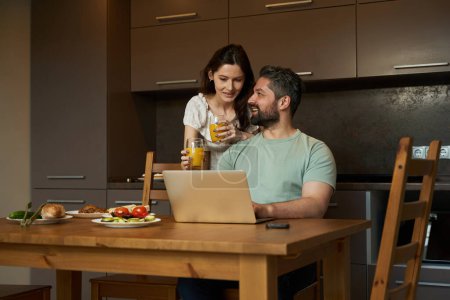 Téléchargez les photos : Homme et femme d'âge moyen communiquent dans une cuisine confortable, une femme traite un homme avec du jus - en image libre de droit