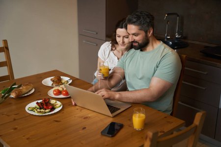 Téléchargez les photos : Homme barbu et une femme mignonne communiquent dans une cuisine confortable, mâle à une table avec un ordinateur portable - en image libre de droit