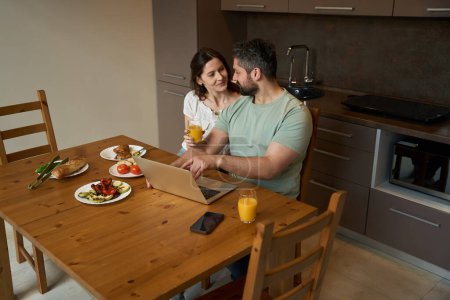 Téléchargez les photos : Mari barbu et une femme douce communiquer dans une cuisine confortable, mâle à une table avec un ordinateur portable - en image libre de droit