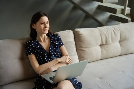 Téléchargez les photos : Femme en robe à pois se trouve sur le canapé à la maison avec un ordinateur portable sur ses genoux, intérieur intérieur moderne - en image libre de droit