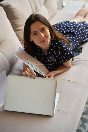 Foto de Señora en un vestido de lunares se encuentra en el sofá con un ordenador portátil y documentos, ella trabaja desde casa - Imagen libre de derechos