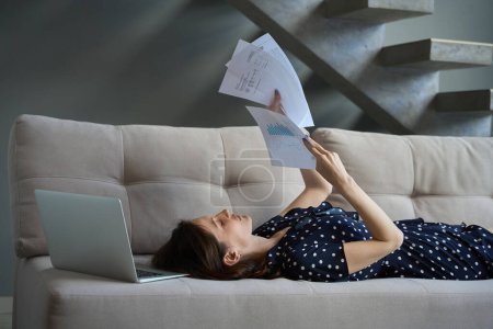 Photo for Brunette in a polka-dot dress lies on the sofa and studies working documents, there is a laptop nearby - Royalty Free Image