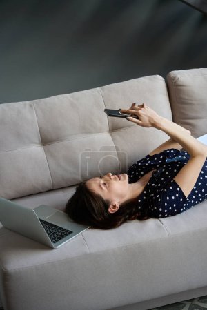 Foto de Morena de pelo largo se encuentra en un sofá suave, tiene un teléfono móvil en sus manos, junto a un ordenador portátil - Imagen libre de derechos