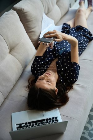 Photo for Beautiful woman communicates online on a mobile phone, next to a laptop, she lies on the couch - Royalty Free Image