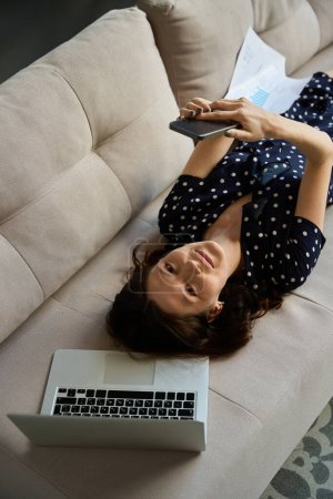 Photo for Female communicates online on a mobile phone, next to a laptop, she is located on the couch - Royalty Free Image