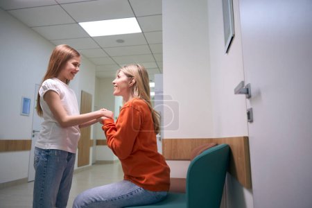 Téléchargez les photos : Mère et fille gaies sont dans un couloir lumineux de l'hôpital, une femme tient les mains des filles - en image libre de droit