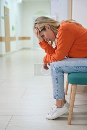 Foto de Rubia femenina trastornada se sienta en un pasillo del hospital, una mujer en un suéter naranja brillante - Imagen libre de derechos