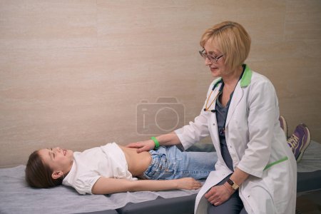 Téléchargez les photos : Femme en robe médicale examine le ventre des filles, l'enfant se trouve sur un canapé médical - en image libre de droit