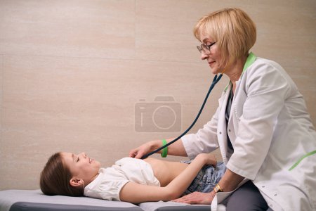 Foto de Pediatra en la recepción escucha a la niña con un statoscopio, el niño se acuesta en un sofá médico - Imagen libre de derechos