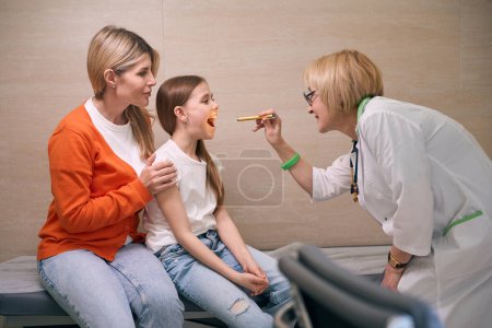 Foto de Pediatra femenina en la clínica examina la garganta de una pequeña paciente, junto a la madre del niño - Imagen libre de derechos