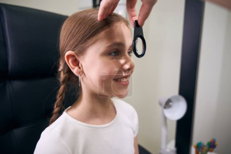 Téléchargez les photos : Spécialiste de la clinique vérifie la vue d'une jeune fille souriante, un ophtalmologiste utilise un outil spécial - en image libre de droit
