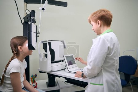 Photo for Girl with two pigtails is in a clinic at an appointment with an ophthalmologist, a woman uses special equipment - Royalty Free Image