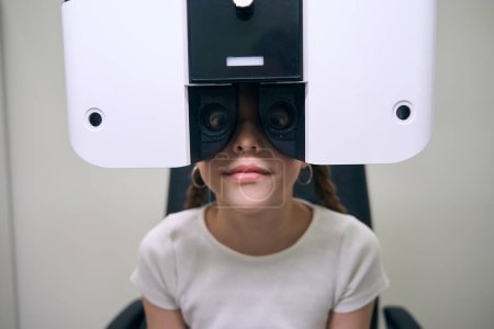 Photo for Young female patient sits in front of a special apparatus for diagnosing vision, a girl in casual clothes - Royalty Free Image