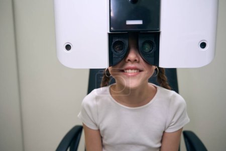 Téléchargez les photos : Fille gaie assis devant un appareil spécial pour diagnostiquer la vision, l'enfant a deux nattes - en image libre de droit