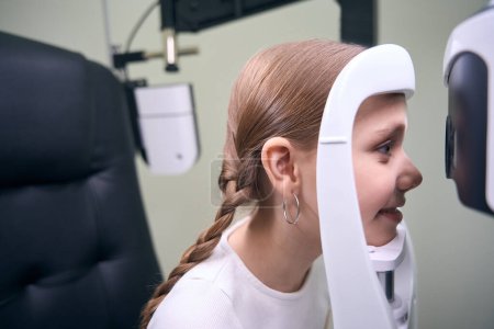 Photo for Cute girl sits in front of a special apparatus for diagnosing vision, the girl has two pigtails - Royalty Free Image