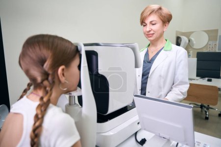 Téléchargez les photos : Femme en robe médicale examine les yeux d'un jeune patient sur un appareil spécial, le médecin utilise un équipement moderne - en image libre de droit