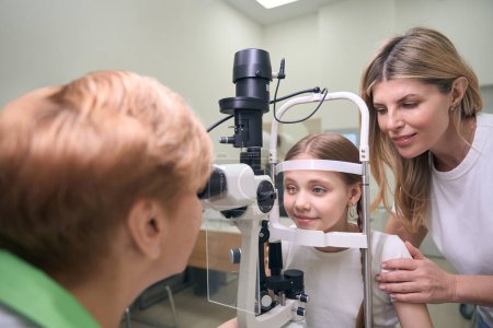 Photo for Girl sits in front of a diagnostic ophthalmological apparatus, next to her mother and doctor - Royalty Free Image
