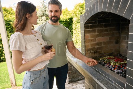 Téléchargez les photos : Un homme souriant cuisine des légumes sur des charbons dans un grand four, à côté de sa femme avec un verre de vin - en image libre de droit