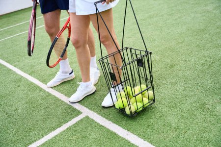 Téléchargez les photos : Partenaires de tennis en vêtements de sport marchent le long du court de tennis, une femme a un panier avec des balles de tennis - en image libre de droit