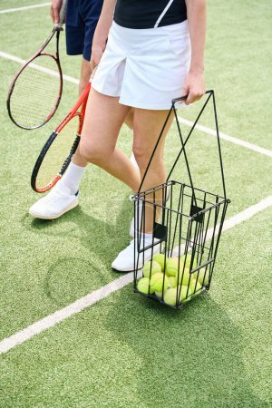 Foto de Los cónyuges caminan con raquetas y una canasta de pelotas en la cancha de tenis, personas en zapatos deportivos cómodos - Imagen libre de derechos